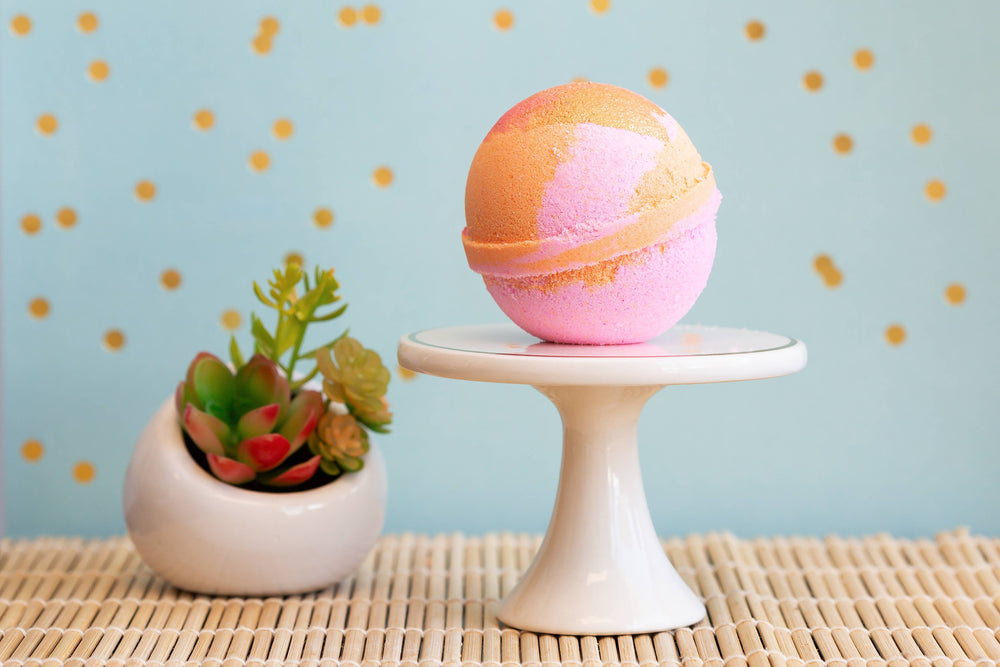 Pink and orange bath bomb on a small white stand with a plant against a light blue background with gold polka dots.